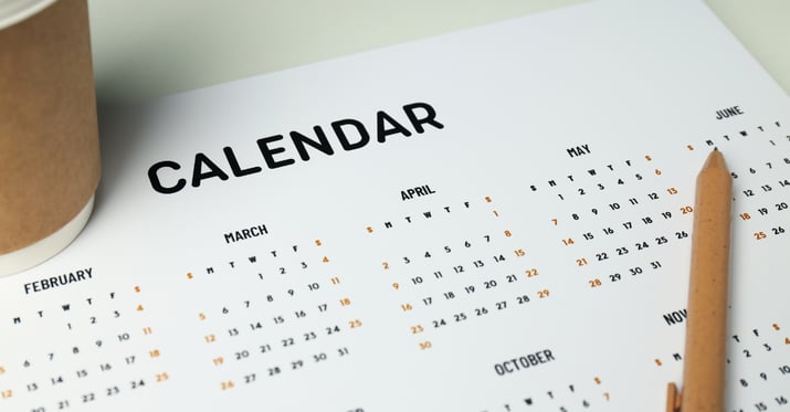 A photo of a to-go coffee laying next to a calendar on a table with a pencil next to the calendar.